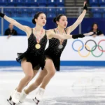 Two figure skaters performing synchronized double axel on Olympic ice with gold medal glinting and scorecard visible