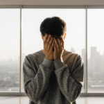 Person holding hands over health concerns while standing with city skyline visible through window wearing affordable fashion