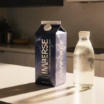 Beyond Immerse recyclable carton stands on kitchen counter with glass bottle of protein drink and golden light
