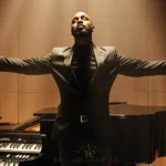 Richard Smallwood stands with arms outstretched beside a grand piano and gospel instruments looking toward a golden ceiling.