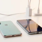 Two smartphones iPhone Air and Galaxy S25 Edge sit side by side on a white desk with soft warm light and a charging cable nea