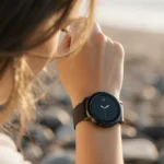 Woman wearing modern smartwatch with circular face and minimalist interface showing natural light and blurred beach stones