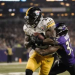 Steelers receiver celebrating touchdown with defender in end zone under golden lights and blurred stadium logo in background