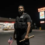 Christian holds an American flag with a baseball bat against his leg in a dim parking lot near a Burger King sign.