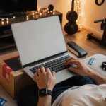 Young adult unwraps tech gadgets revealing a shiny Apple M4 laptop with gift cards and fitness trackers on a couch in a gym.