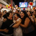 Venezuelan immigrants hugging and cheering at Preciosa Market with TV showing Maduro capture surrounded by floral decorations