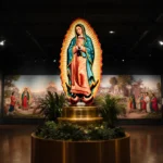 La Virgen de Guadalupe stands on golden pedestal with lush greenery and colorful textiles glowing under warm light