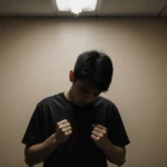 Young college student stands with head bowed and clenched fists under flickering light showing wrongful deportation despair